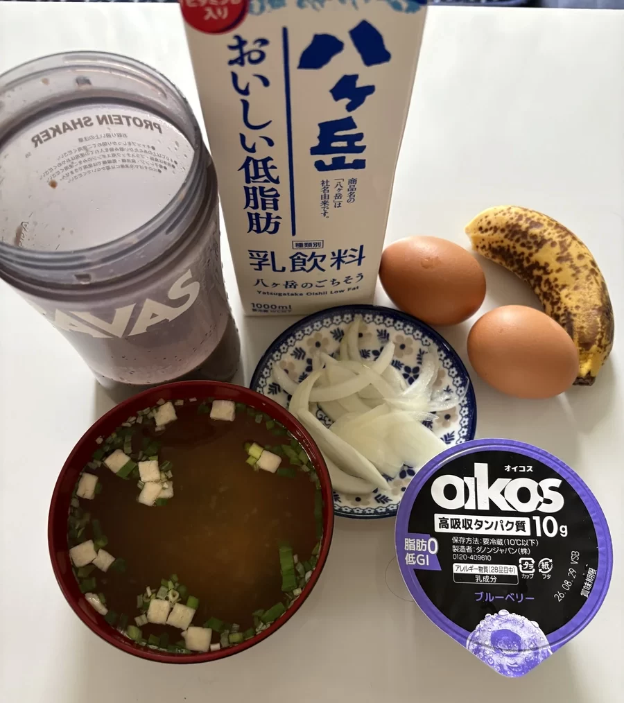 朝ごはん一式。バナナ、ゆで卵2個、ヨーグルト、玉ねぎ、味噌汁、低脂肪牛乳と並んで、プロテインを入れたシェイカーが置いてある。